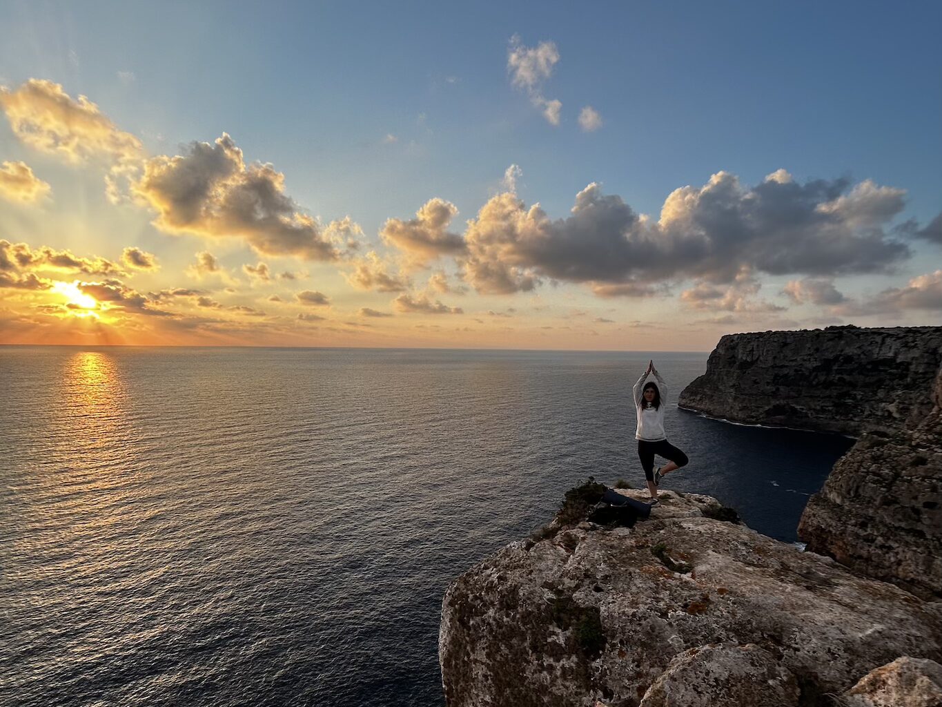 Femme en posture de l'arbre (Vrikshasana) sur une falaise de Formentera au lever du soleil, face à la mer.