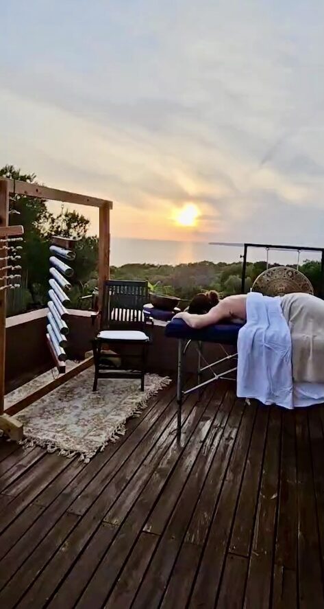 Soin énergétique et massage sur une terrasse en bois à Formentera, avec carillon éolien et gong face au coucher du soleil.